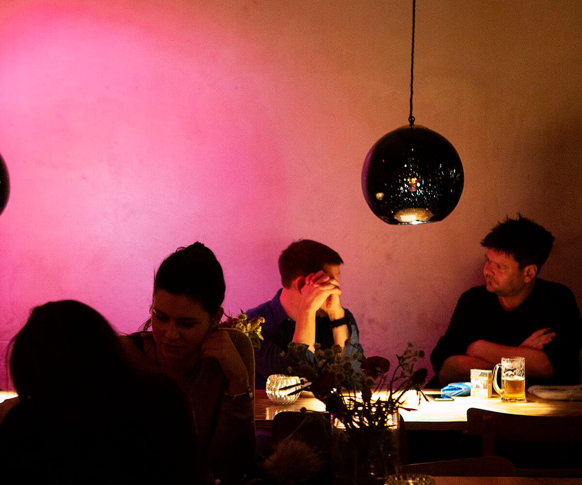 Dunkel beleuchtete Bar mit Gästen, die an Tischen sitzen; pinke Wandbeleuchtung und hängende Lampen.