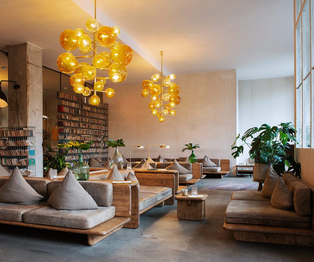 Moderne Hotel-Lobby mit Holzsofas, Pflanzen und auffälligen, runden, gelben Lampen. Bücherregal im Hintergrund.