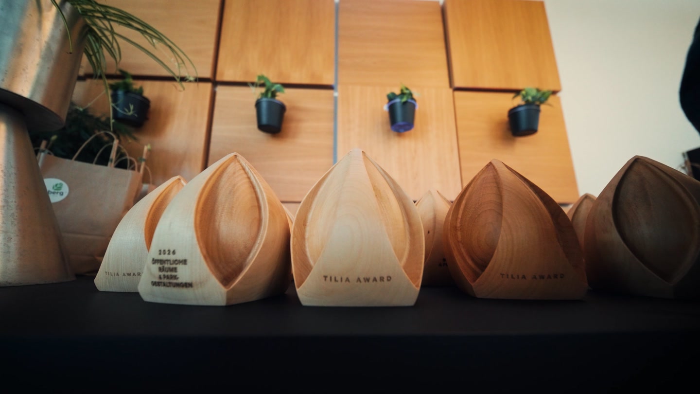 Eine Reihe der hölzernen Tilia-Award-Skulpturen auf einem Tisch, dahinter Setzlinge in kleinen Töpfen an einer Holzwand.
